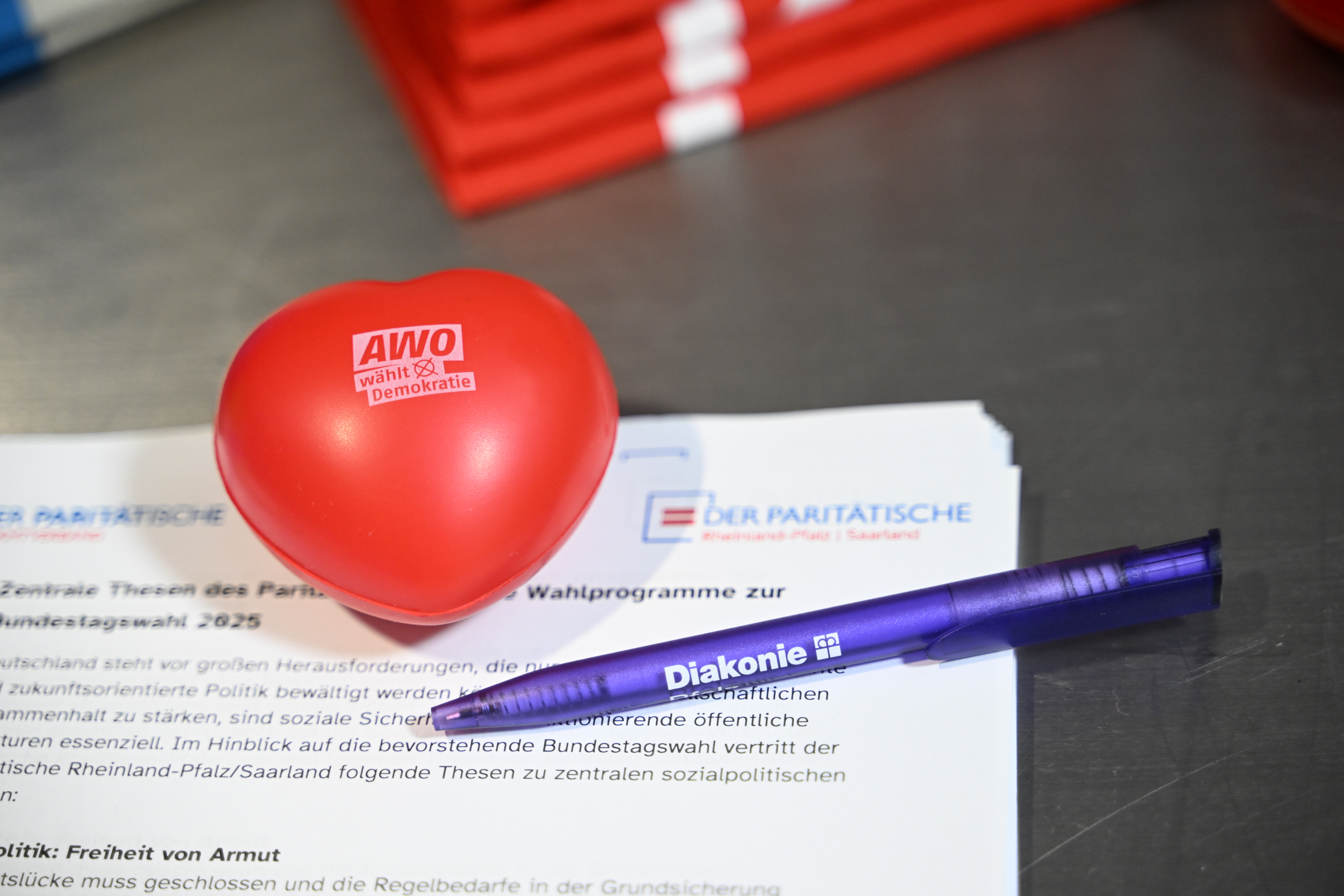Roter Stressball in Form eines Herzens mit dem Aufdruck „AWO wählt Demokratie“, ein Dokument mit dem Logo des Paritätischen Wohlfahrtsverbands Rheinland-Pfalz und ein Kugelschreiber der Diakonie.